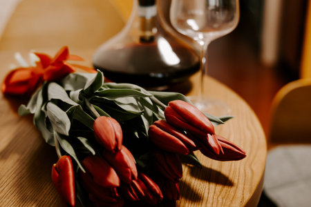 A bouquet of red tulips on a wooden table with a wine decanter and glass in the background. Moody vintage still life with soft lighting and film grain. Ideal for romantic and celebration designs.の写真素材