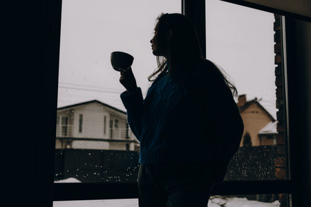 Silhouette of a young woman with a cup of coffee looking out of a window at a snowy backyard. Atmospheric moody indoor scene of solitude and winter comfort. Minimalist lifestyle photography backgroundの写真素材