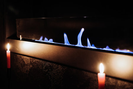 Modern bioethanol fireplace with vibrant blue flames and lit candles. Moody dark aesthetic with warm...