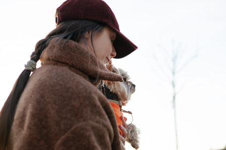 Candid portrait of a woman in a maroon cap and brown fleece jacket holding a small dog in an orange...