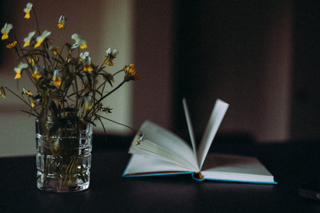 Still life featuring an open book and a bouquet of wildflowers in a glass vase on a dark table. Cozy interior scene symbolizing reading, leisure, quiet relaxation, home comfort and mindful living.