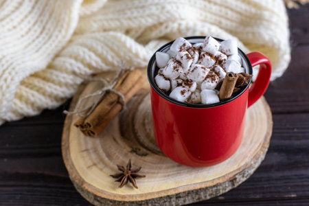 Hot chocolate with marshmallows, cinnamon, star anise - a winter hot spicy drink in a red cup on a wooden cut, with a white knitted scarf on a wooden backgroundの写真素材