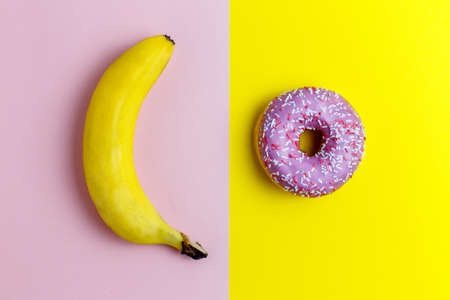 Healthy vs unhealthy food, dieting concept. Pink donuts, doughnut with sweet sprinkles on yellow and a banana isolated on pink color backgroundの写真素材