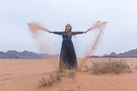 A young woman with blond hair in a long dark blue dress is playing with sand. Wadi Rum Desert Jordanの写真素材