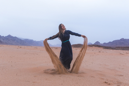 A young woman with blond hair in a long dark blue dress is playing with sand. Wadi Rum Desert Jordanの写真素材