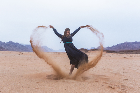 A young woman with blond hair in a long dark blue dress is playing with sand. Wadi Rum Desert Jordanの写真素材