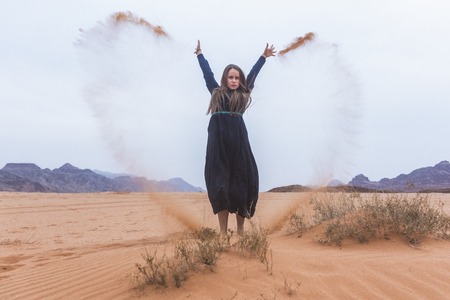 A young woman with blond hair in a long dark blue dress is playing with sand. Wadi Rum Desert Jordanの写真素材