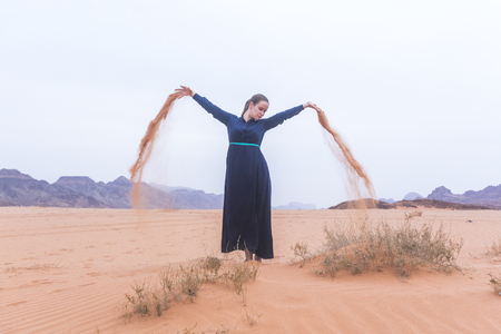 A young woman with blond hair in a long dark blue dress is playing with sand. Wadi Rum Desert Jordanの写真素材
