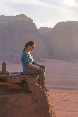 the girl-traveler sits on a stone ledge against the backdrop of the mountains in the Wadi Wadi desert at sunsetの写真素材