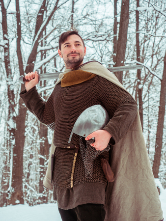 Medieval viking warrior in chain mail and helmet holds a sword in the winter forestの写真素材