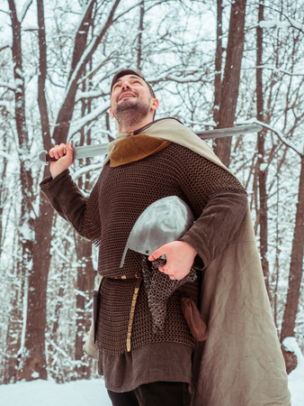 Medieval viking warrior in chain mail and helmet holds a sword in the winter forestの写真素材