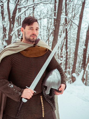 Medieval viking warrior in chain mail and helmet holds a sword in the winter forestの写真素材