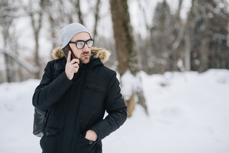 Profile of attractive happy young man with beard, phone standing in winter city. Glasses, jacket,...