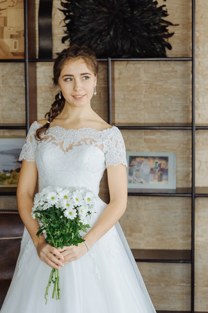 Beautiful bride in white wedding dress standing in her room and near bookselfs with flowers.の写真素材