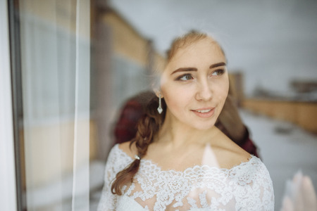 Beautiful bride in white wedding dress standing in her bedroom and looking in windowの写真素材