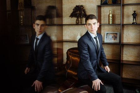 The handsome groom sitting in modern hall near bookselfs. Man looking on his wedding day and reflex mirror.の写真素材