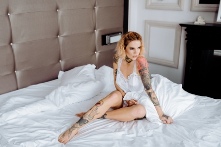 Beautiful bride with tattoo in lingerie showing sexy pose, sitting at bed over her bedroom. Color. Sexual memories.の写真素材