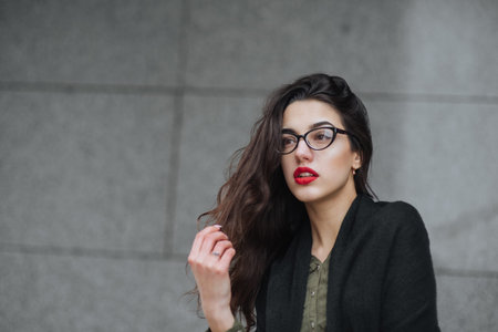Fashion consept: beatiful young girl with long hair, glasses, red lips standing near modern wall wearing in green suit and grey jeans. She wanted talking on phone. Colorの写真素材