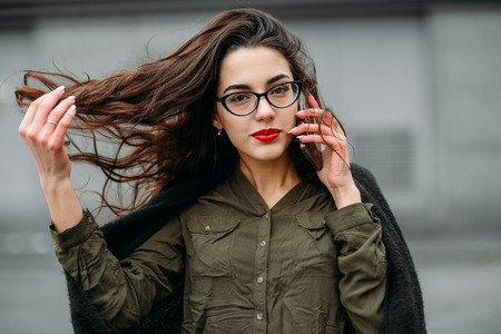 Fashion consept: beatiful young girl with long hair, glasses, red lips standing near modern wall wearing in green suit and grey jeans. She talking on phone. Colorの写真素材
