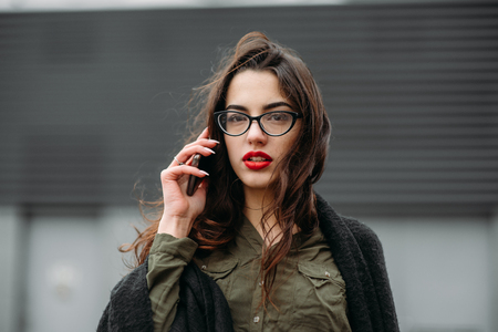 Fashion consept: beatiful young girl with long hair, glasses, red lips standing near modern wall wearing in green suit and grey jeans. She talking on phone. Colorの写真素材