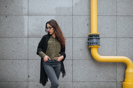 Fashion consept: beatiful young girl with long hair, glasses, red lips standing near modern wall...