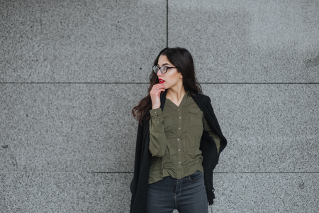 Fashion consept: beatiful young girl with long hair, glasses, red lips standing near modern wall wearing in green suit and grey jeans. She wanted talking on phone. Colorの写真素材