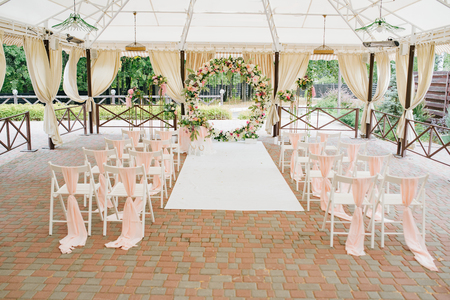 Beautiful wedding set up. Area of the wedding ceremony. Round arch, white chairs decorated with flowers, greenery. Cute, trendy rustic decor. Part of the festive decor, floral arrangement.の写真素材