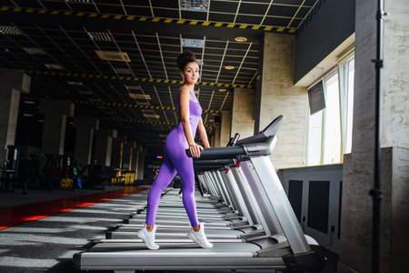 Sport world. Full-length side view of handsome woman in violet sportswear running on a treadmill in...