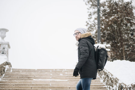 Profile of attractive happy young man with beard standing in winter city. Glasses, jacket, hat, bagpack. Color.の写真素材