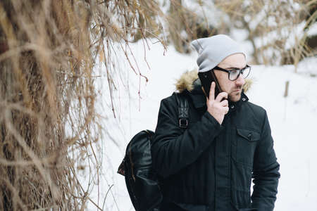 Profile of attractive happy young man with beard standing in winter urban. Glasses, jacket, hat, phone. Color.の写真素材