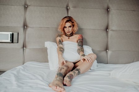 Portrait of a happy beautiful girl with tattoos. The woman is sitting in bed and covers his body with a pillow.の写真素材