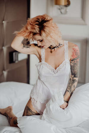 Portrait of a happy beautiful girl with tattoos. The woman is sitting in bed and touch her curly hair.の写真素材