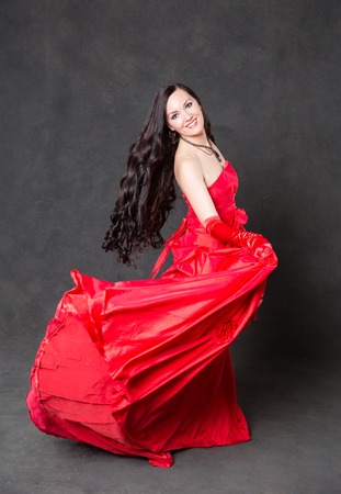 Latino Woman with long hair  in red waving dress dancing  in action with flying fabric on dark grey backgroundの写真素材