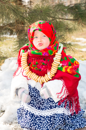 Child girl in Russian pavloposadskie folk scarf on head with floral print and with  bunch of bagels on background of snow. Portrait of girl dressed in Russian styleの写真素材