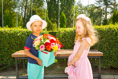 Black african american boy kid gives flowers to girl child on birthday. Little adorable children in park. Childhood and love.の写真素材