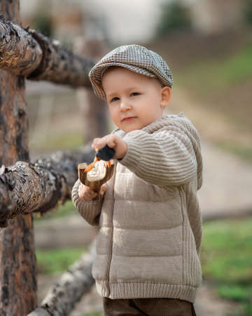 Bully boy kid with a slingshot aims at someone near a fence in the village outdoors. Rustic barefoot child boy with a hat shoots a slingshot.の写真素材