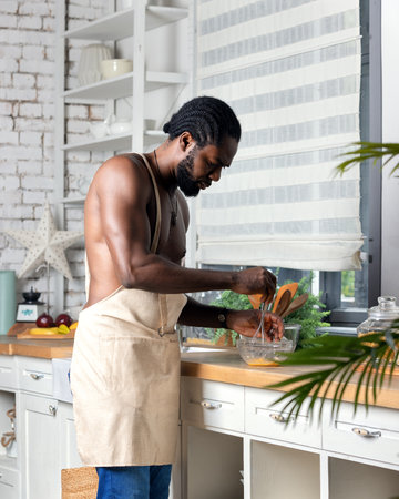 Black man cooking breakfast or lunch on kitchen at home. African American man wearing an apron preparing fresh. Raw ingredients on table. Healthy food concept. Family communicating, cooking and enjoying weekend timeの写真素材