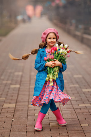 Beautiful girl kid with spring pink tulips flowers bouquet at city street. Portrait of pretty child in park outdoors.の写真素材