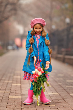 Beautiful girl kid with spring pink tulips flowers bouquet at city street. Portrait of pretty child in park outdoors.の写真素材