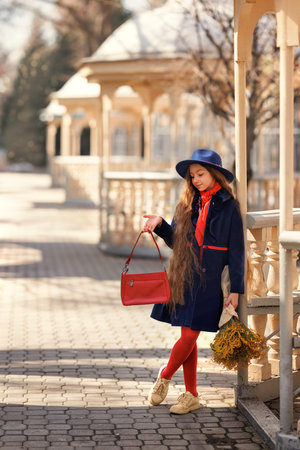 Beautiful girl kid in hat with spring yellow mimosa flowers bouquet at city street. Portrait of pretty child with long hair in park outdoors.の写真素材