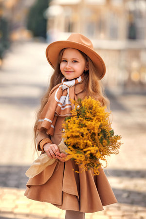 Beautiful girl kid in hat with spring yellow mimosa flowers bouquet at city street. Portrait of pretty child with long hair in park outdoors.の写真素材