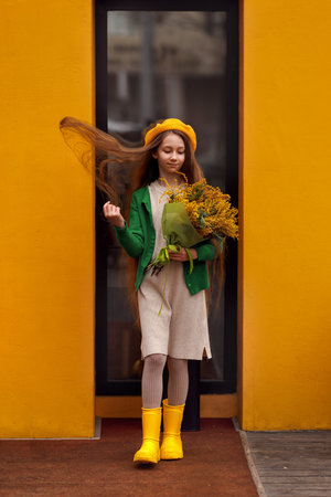 Beautiful girl kid in beret with spring yellow mimosa flowers bouquet at city street. Portrait of pretty child with long hair in park outdoors.の写真素材