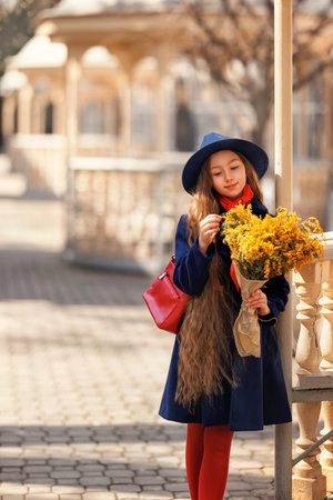 Beautiful girl kid in hat with spring yellow mimosa flowers bouquet at city street. Portrait of pretty child with long hair in park outdoors.の写真素材