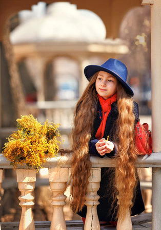 Beautiful girl kid in hat with spring yellow mimosa flowers bouquet at city street. Portrait of pretty child with long hair in park outdoors.の写真素材
