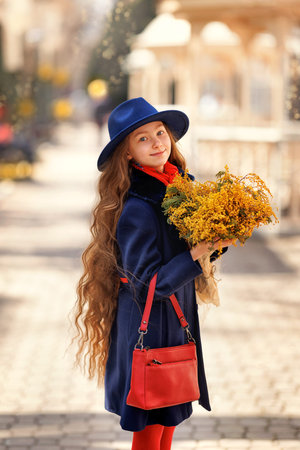 Beautiful girl kid in hat with spring yellow mimosa flowers bouquet at city street. Portrait of pretty child with long hair in park outdoors.の写真素材