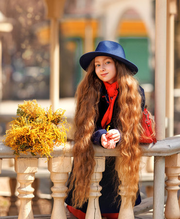 Beautiful girl kid in hat with spring yellow mimosa flowers bouquet at city street. Portrait of pretty child with long hair in park outdoors.の写真素材