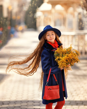 Beautiful girl kid in hat with spring yellow mimosa flowers bouquet at city street. Portrait of pretty child with long hair in park outdoors.の写真素材