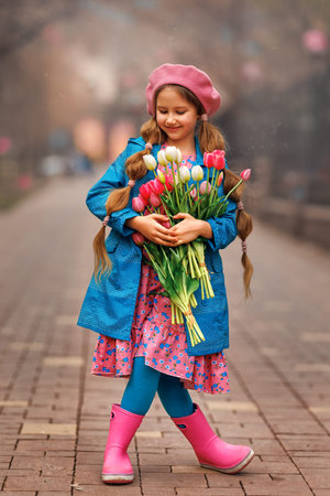 Beautiful girl kid with spring pink tulips flowers bouquet at city street. Portrait of pretty child in park outdoors.の写真素材
