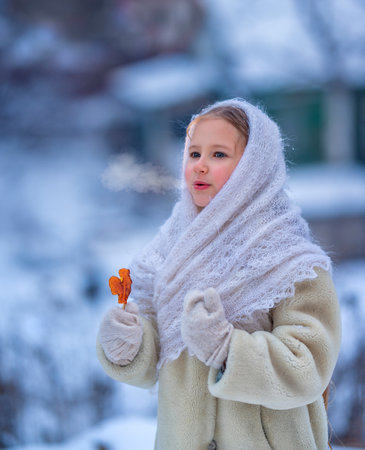 Portrait of a beautiful child girl in a downy shawl with a lollipop on a snowy evening in winter outside.の写真素材