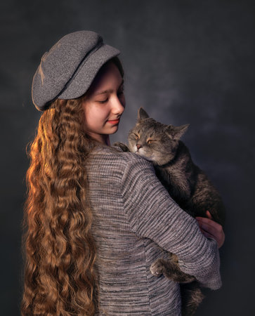 Smiling girl kid with long curly hair in vintage clothes holding grey cat in studio portrait. The cat licks its lips playfullyの写真素材